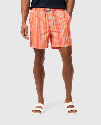 A person wearing Psycho Bunny MENS ANTON ALL OVER PRINT SWIM TRUNK - B6W542C200 with a blue drawstring is standing. They are also wearing white sandals. The background is plain and light-colored.