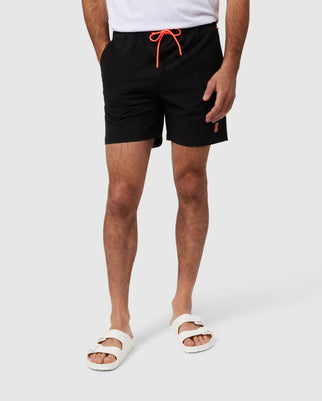 A person wearing black shorts with an orange drawstring, a white T-shirt partially visible, and white slip-on sandals is standing against a plain, light gray background. Their hands are in the pockets of the Psycho Bunny MENS LANCASTER HYDROCHROMIC SWIM TRUNK - B6W632A2PO featuring a hidden Bunny logo.