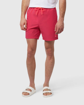 A person stands against a plain background wearing a white t-shirt and vibrant red quick-drying textile shorts with a drawstring. The person is also wearing white slide sandals. The MENS LANCASTER HYDROCHROMIC SWIM - B6W632A2PO by Psycho Bunny features a small silicone Bunny patch on the left thigh. The person's arms are relaxed at their sides.