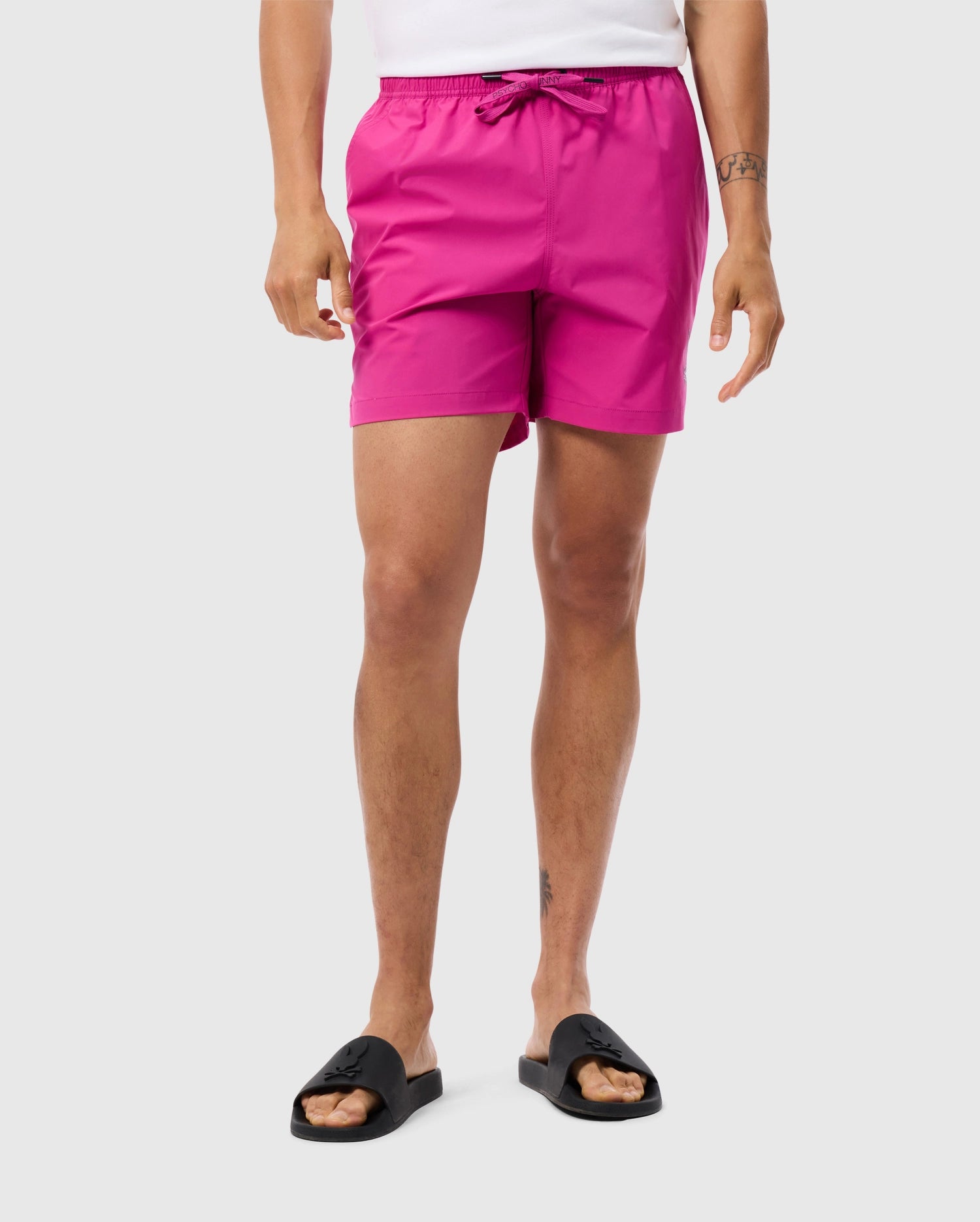 A person is standing against a plain background wearing a white shirt, MENS PARKER HYDROCHROMIC SWIM TRUNK - B6W646C200 by Psycho Bunny featuring a drawstring waistband and bunny pattern, black open-toe sandals, and showcasing tattoos on their left wrist and lower right leg. The person is standing with their arms relaxed by their sides.