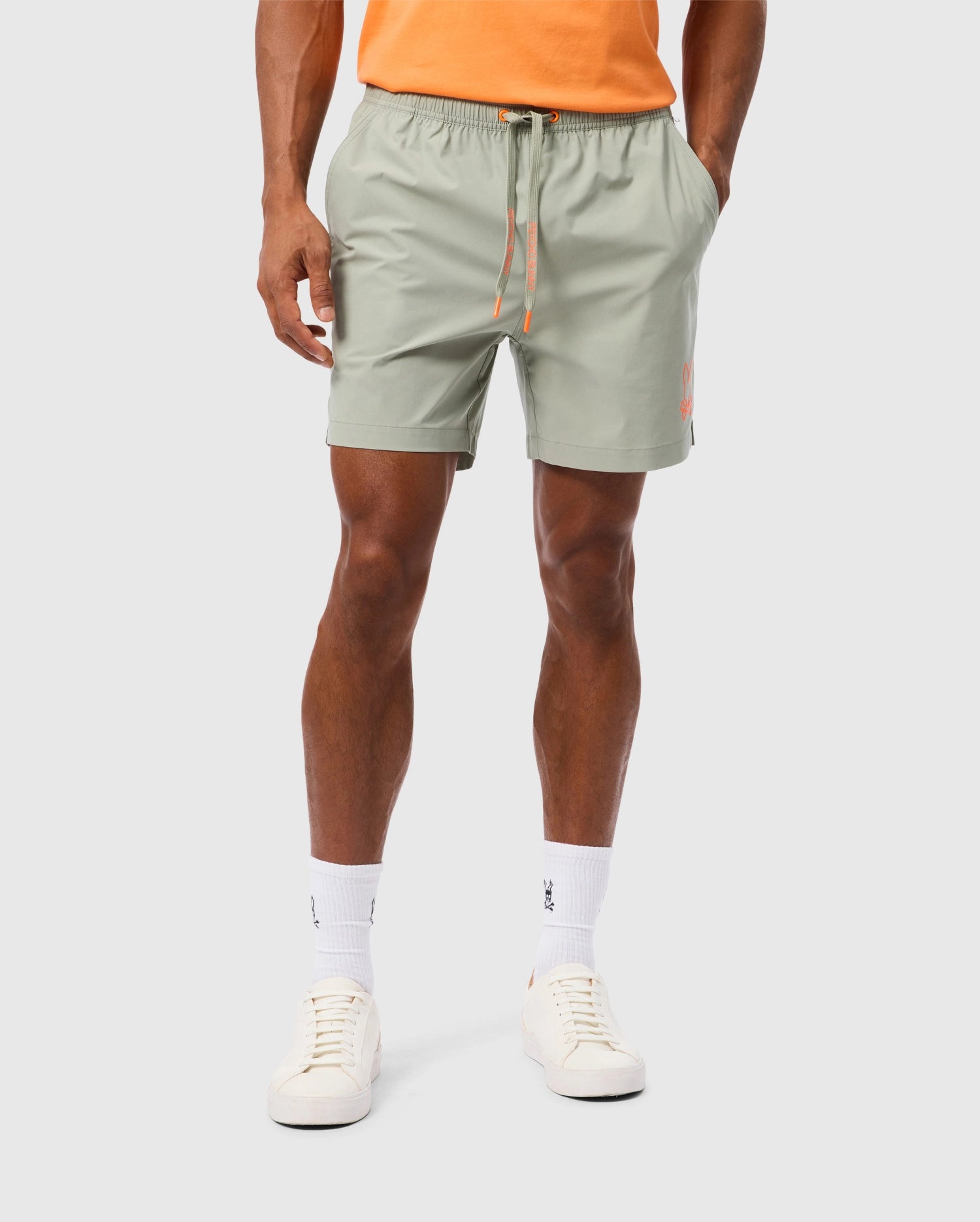 A person wearing light green Psycho Bunny MENS PARKER HYDROCHROMIC SWIM TRUNK - B6W646C200 with an orange drawstring and hands in pockets, showcasing white socks paired with white sneakers. The person is also wearing an orange shirt, the photo highlighting a waist-to-knee view against a plain background. The quick-dry material of the swim trunk ensures comfort.