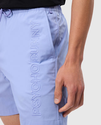 Close-up of a person's hip and hand wearing MENS MELVIN SWIM TRUNK - B6W665C200 in light purple with Psycho Bunny embroidered vertically on the side. The person is also carrying a waterproof bag and wearing a black shirt.