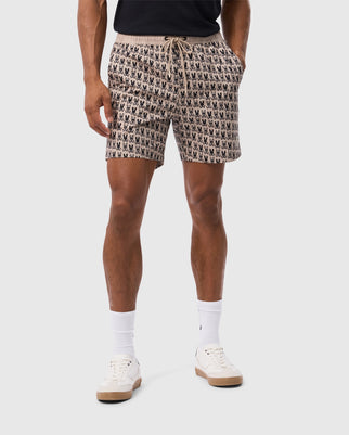 A person is wearing the Psycho Bunny MENS TILDEN PRINT SWIM TRUNK - B6W729D200, featuring small black designs on a beige background, paired with a black T-shirt, white socks, and white sneakers. The individual stands against a plain light-colored backdrop.