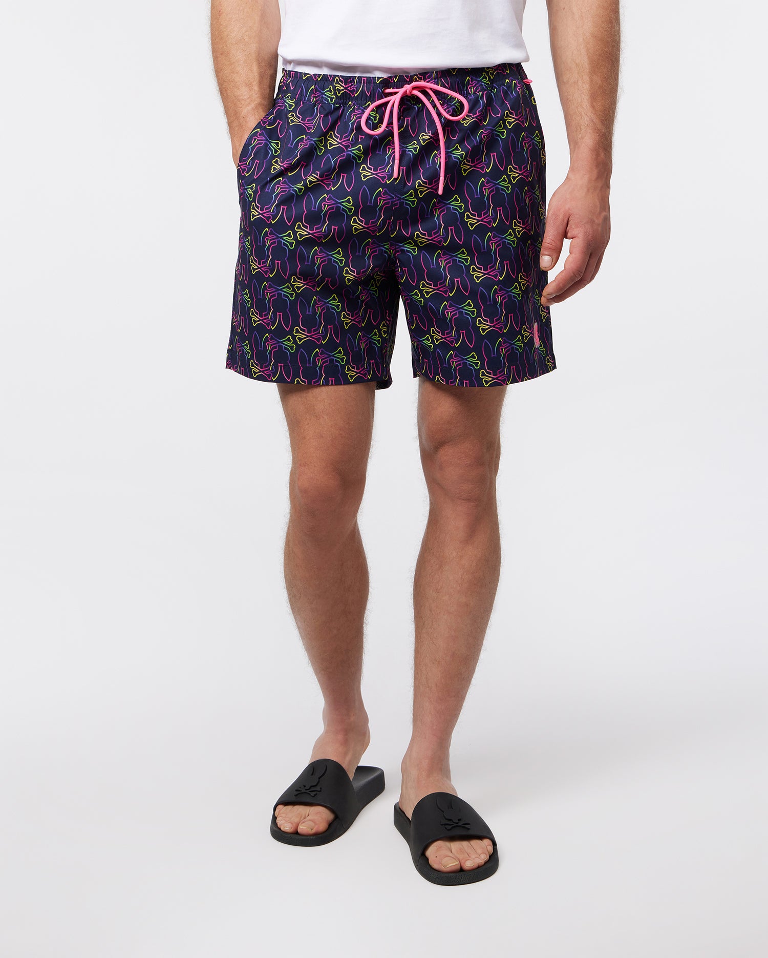 A person is wearing Psycho Bunny MENS COLTON PRINTED SWIM - B6W763A2PO with a brightly-colored abstract pattern and a pink drawstring. They are also wearing black slide sandals. The background is plain white. The person's hands are at their sides and only their lower body is visible.