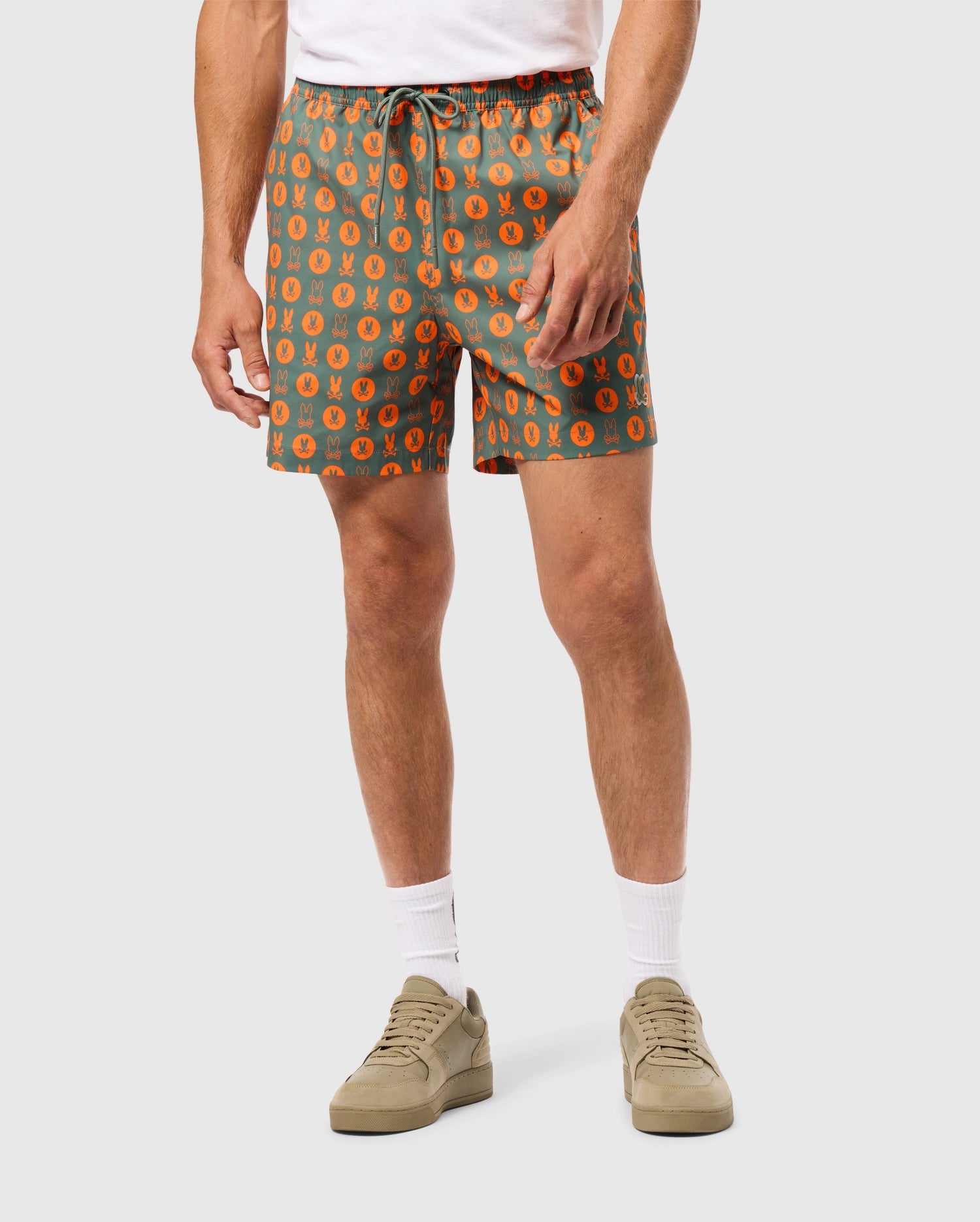 A person is wearing a pair of MENS ARNOLD PRINT SWIM TRUNK - B6W820D200 by Psycho Bunny featuring a green color with an orange pattern and a white drawstring, accompanied by white socks and light brown shoes. The quick-dry material ensures they stay comfortable all day. The background is plain light gray. The person is shown from the waist down.