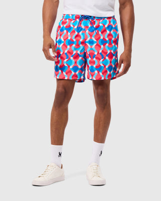 A person is wearing vibrant red, white, and blue geometric-pattern Psycho Bunny MENS WALTER LIGHTWEIGHT PRINT SWIM TRUNK - B6W863D200 made from 100% polyester with a drawstring, white crew socks featuring a small black logo, and white sneakers. The background is plain white.