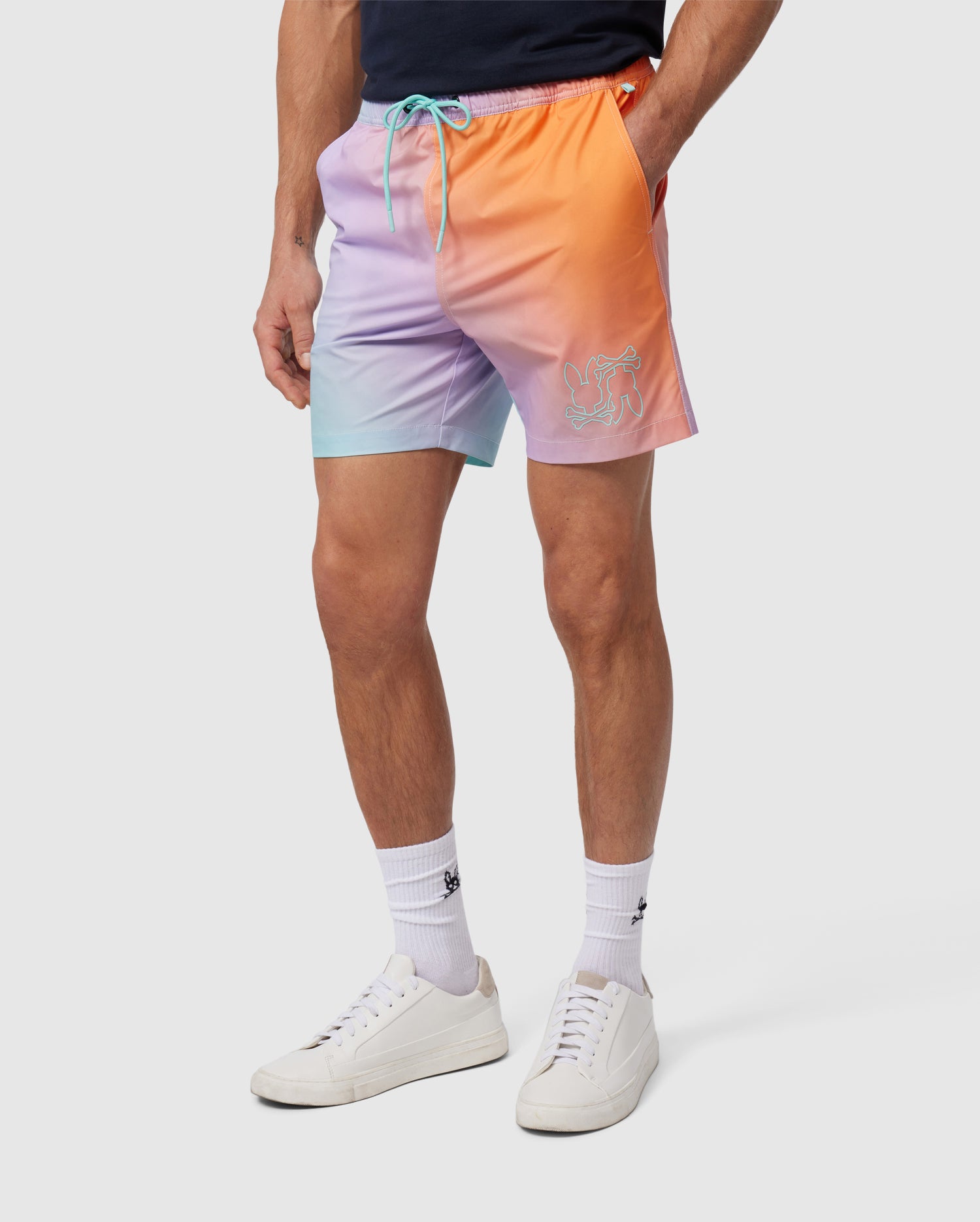 A person wearing a pair of Psycho Bunny MENS WINTON PRINTED SWIM TRUNK - B6W915A2PO with pastel colors of purple, orange, and blue. The trunks feature a small embroidered design on the left leg. They are also wearing white sneakers, white crew socks, and a dark blue t-shirt.