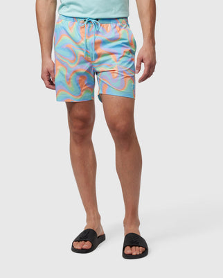 A person standing against a white background wearing Psycho Bunny MENS BLOOMINGTON SWIM TRUNK - B6W941A2PO with a pastel-colored swirling pattern and black slide sandals. Only the lower body from the waist down is visible.