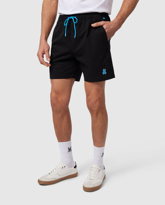 A person is wearing Psycho Bunny's MENS KERMAN HYDROCHROMIC SWIM TRUNK - B6W942A2PO, which are quick-drying black athletic shorts featuring a blue drawstring and a small blue emblem on the left leg. They are also sporting white socks and white sneakers with a light brown sole against a plain white background.