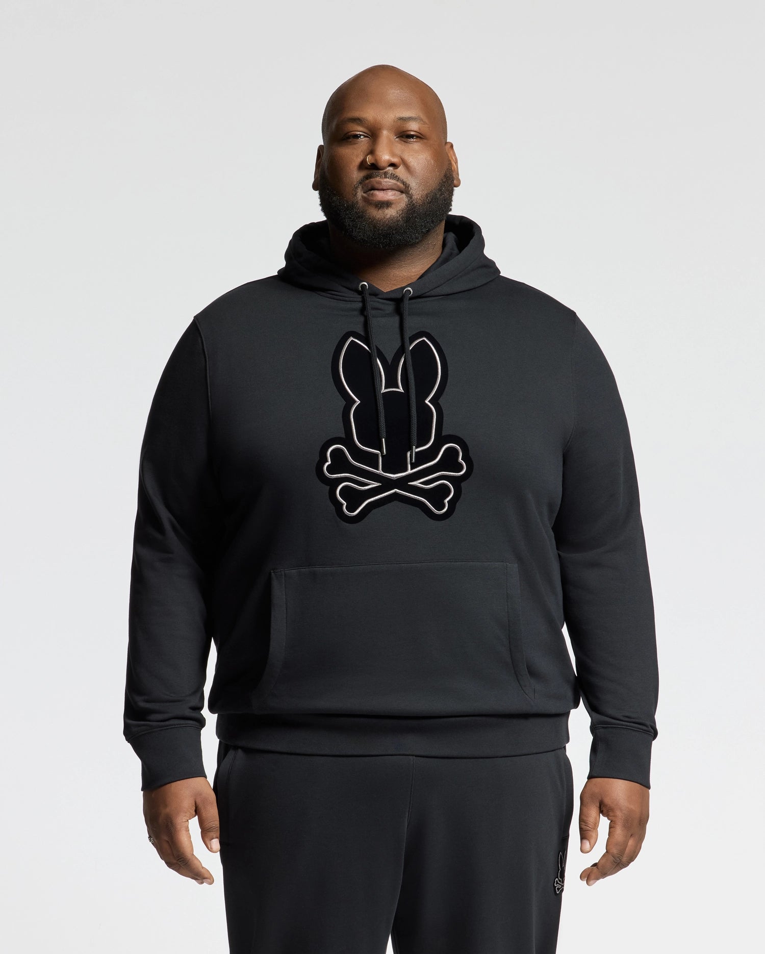 A bearded man confidently stands against a plain background, wearing the Psycho Bunny BIG AND TALL ALEXANDER HOODIE - B9H154E200. This eye-catching black pullover hoodie features a striking bunny and crossbones design on the front. Complemented by matching black pants, it offers a stylish choice perfect for big and tall individuals.