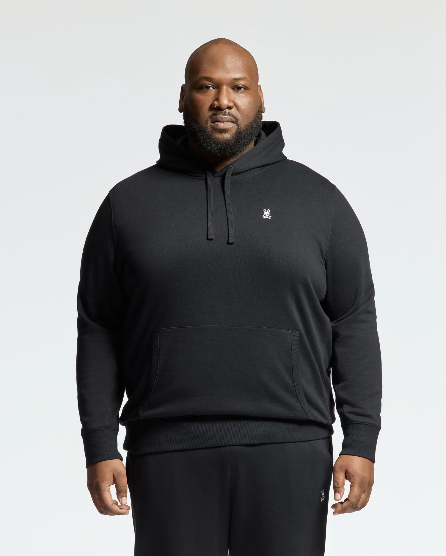 A person stands confidently in a black Psycho Bunny MENS BIG AND TALL LEVY ESSENTIAL FLEECE HOODIE - B9H859D200 and sweatpants against a light background. The 100% cotton hoodie, featuring a small embroidered logo and kangaroo pocket, accentuates their style. They look directly at the camera with hands by their sides.