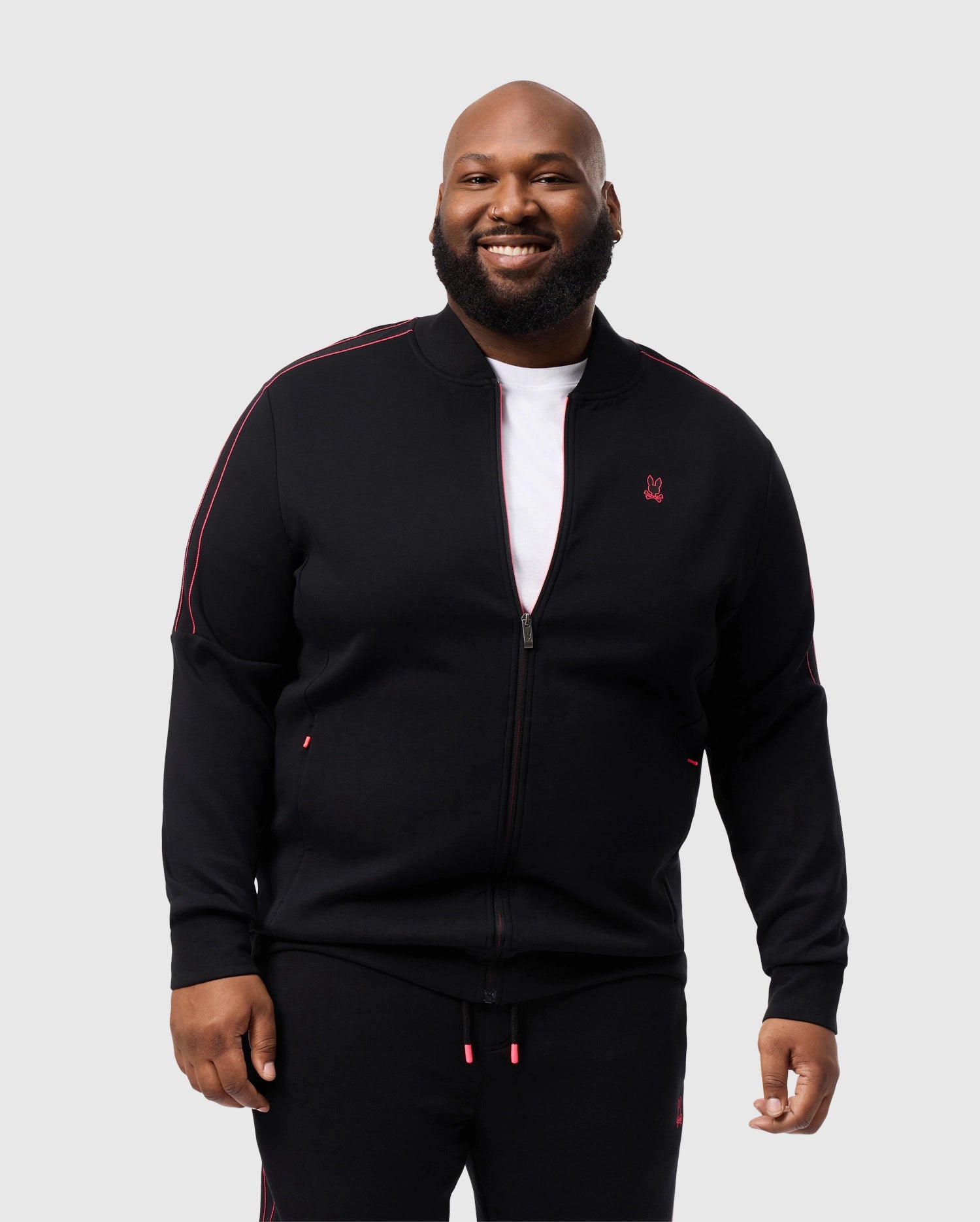 A smiling bearded man wears the Psycho Bunny BIG AND TALL TRAVIS TRACK JACKET (B9J142D200), a black cotton-blend tracksuit with red accents and a zippered front, featuring a silicone emblem. He stands against a plain light gray background.