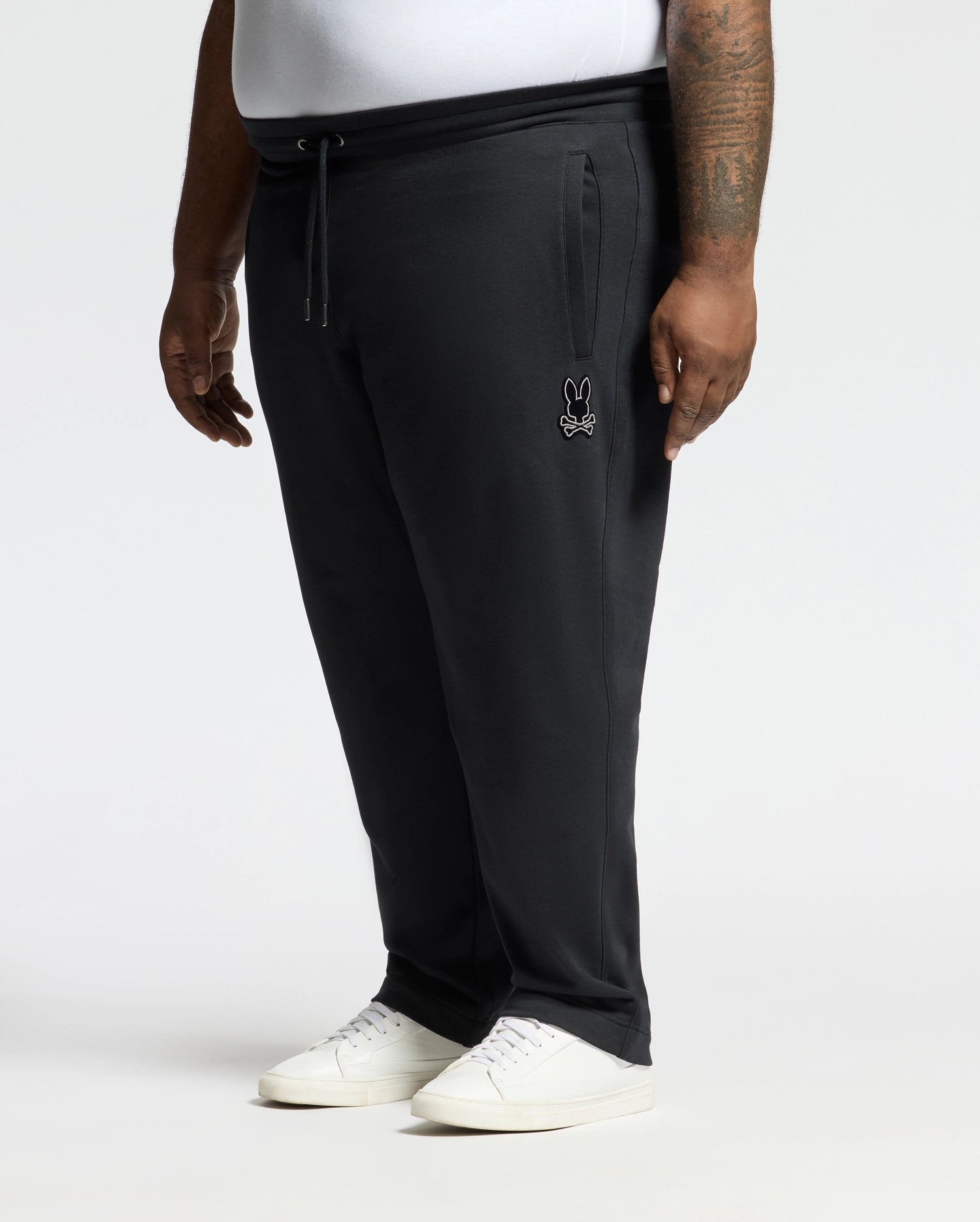 Someone is wearing comfortable black BIG AND TALL ALEXANDER SWEATPANT - B9P155E200 by Psycho Bunny with a small bunny logo near the pocket, paired with a white shirt and sneakers against a plain white background.