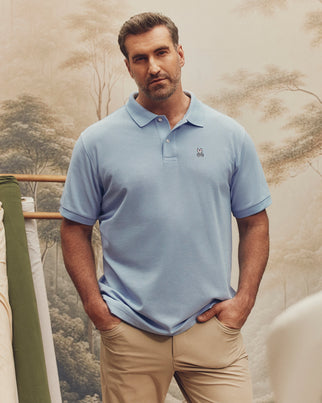 A man stands confidently against nature-themed wallpaper in a light blue Psycho Bunny BIG AND TALL CLASSIC PIQUE POLO - B9K001CRPC and beige pants.