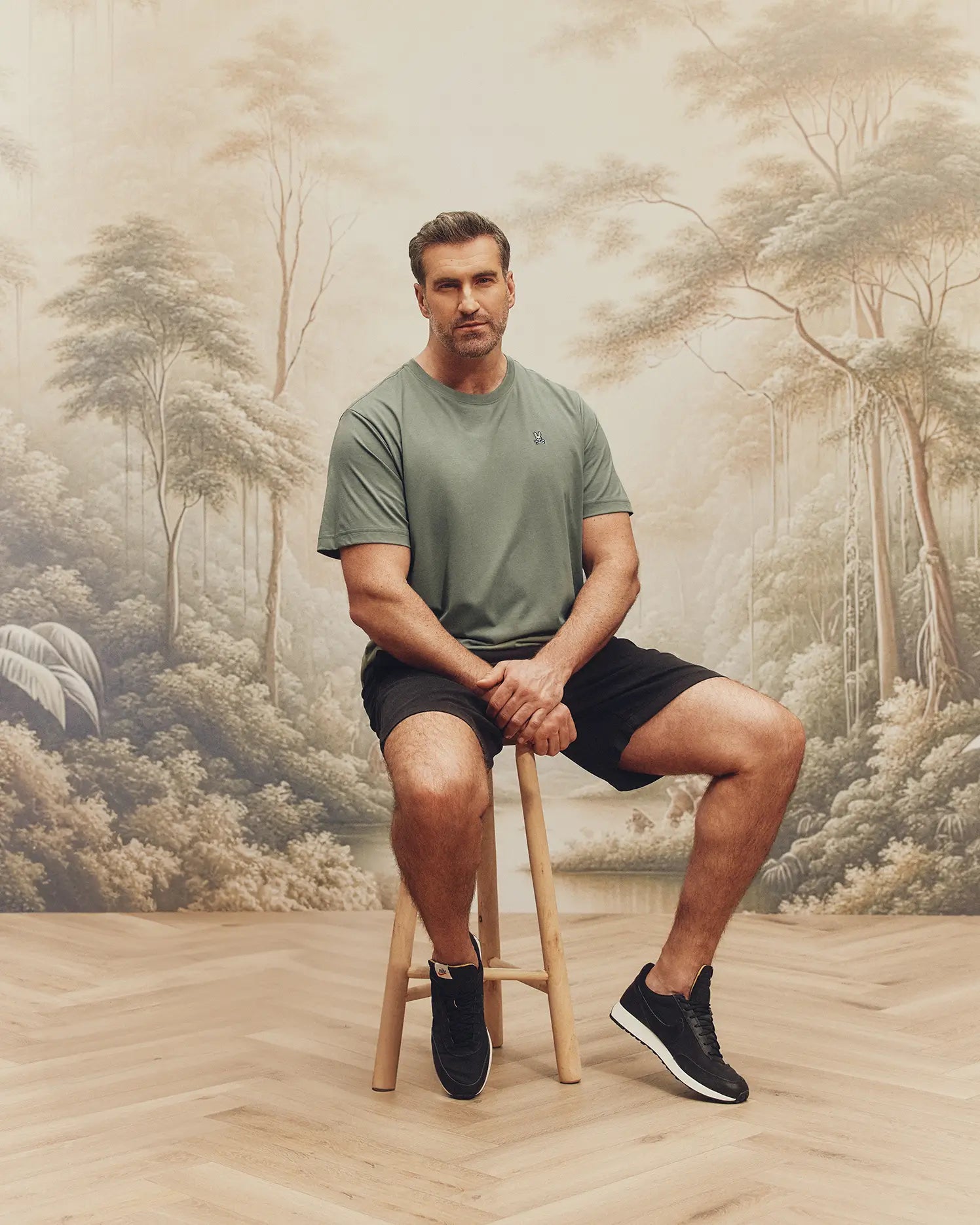 A man sits on a wooden stool before a serene forest mural, wearing a Psycho Bunny Big and Tall Classic Crew Neck Tee (B9U014CRPC) in green, with black shorts and sneakers. Beside him, a basket of cotton sprigs rests on the wood floor, enhancing his casual yet stylish look.