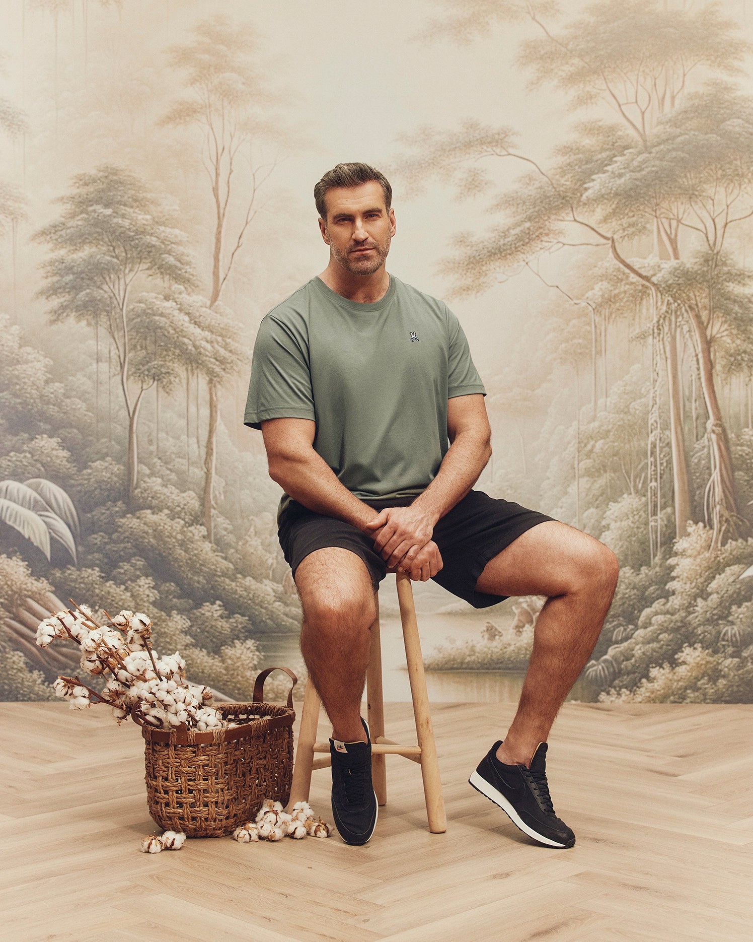 A man sits on a wooden stool before a serene forest mural, wearing a Psycho Bunny Big and Tall Classic Crew Neck Tee (B9U014CRPC) in green, with black shorts and sneakers. Beside him, a basket of cotton sprigs rests on the wood floor, enhancing his casual yet stylish look.