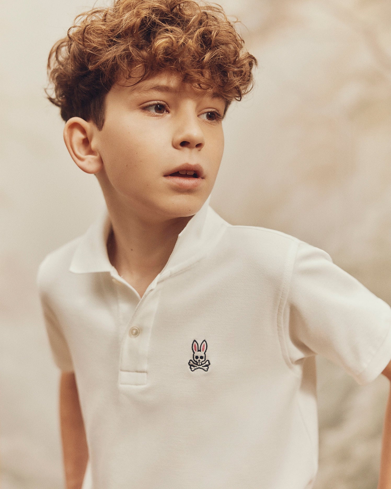 A young boy with curly hair models the Psycho Bunny KIDS CLASSIC PIQUE POLO SHIRT - B0K001CRPC, showcasing its small bunny emblem on the chest. He poses against a soft, neutral background, looking off to the side, ideal for back-to-school outfits.