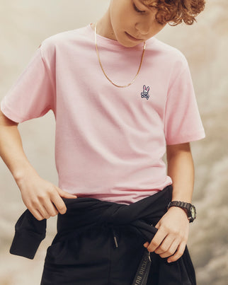 A young person in a pink Psycho Bunny KIDS CLASSIC CREW NECK TEE, made from 100% Pima cotton with an embroidered bunny logo, adjusts a black jacket tied around their waist. With curly hair, a gold chain, and black watch, they're set against a softly blurred backdrop for an effortlessly cool back-to-school look.