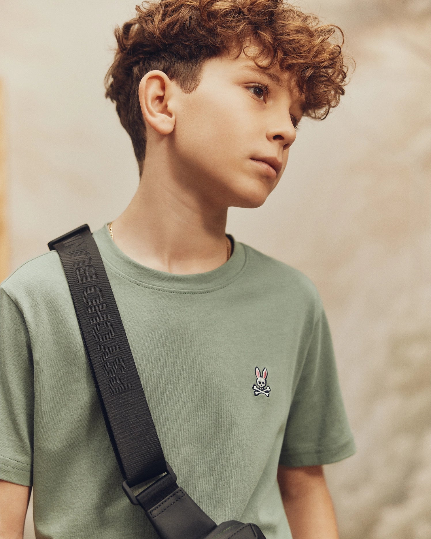 A young boy with curly hair, wearing Psycho Bunny's KIDS CLASSIC CREW NECK TEE in green, 100% Pima cotton, featuring a small rabbit emblem and a black crossbody bag, looks thoughtfully to the side. The softly blurred background evokes perfect back-to-school vibes.