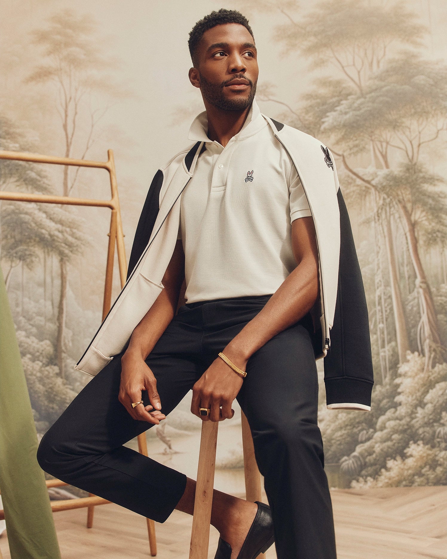 A bearded man sits on a wooden stool wearing a white MENS CLASSIC PIQUE POLO SHIRT - B6K001ARPC by Psycho Bunny, with a black jacket draped over his shoulders and black pants. The nature-themed mural in the background features trees and soft, earthy tones.