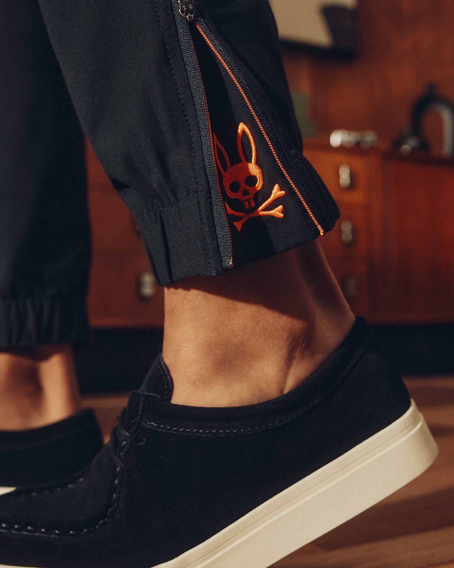 A close-up shows a person wearing Psycho Bunny's MENS MADISON COMMUTER PANT with orange skull and bunny ear embroidery near the ankle and zipper detail. The elastic waistband adds comfort, complemented by black shoes with white soles, against a wooden floor, framed by a vintage cabinet.