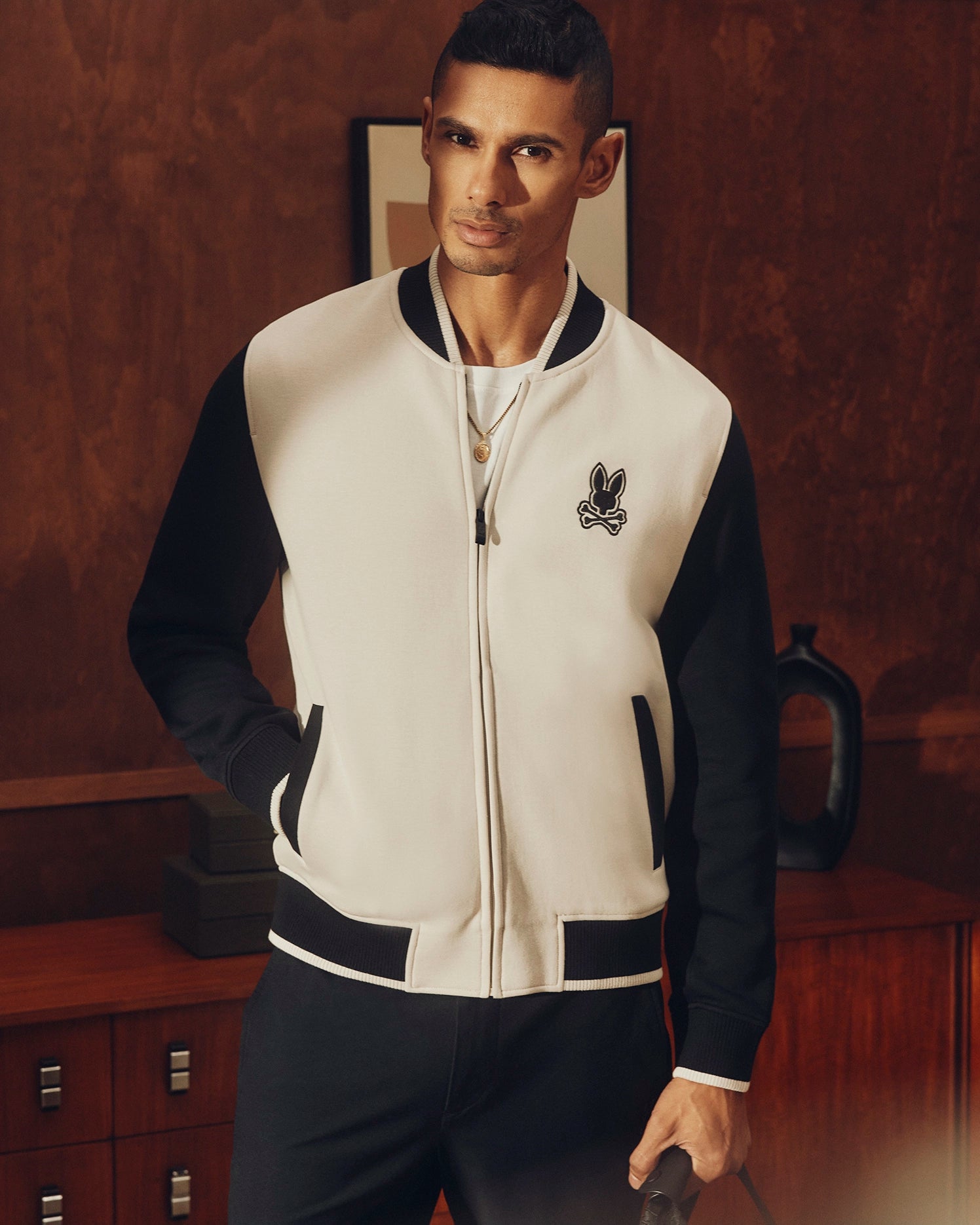 In a room with wooden walls and a cabinet, a man confidently wears the Psycho Bunny MENS CRETE COLOR BLOCK BOMBER - B6J421F200. The black-and-white jacket features an elegant chest embroidery and classic cut, enhancing his poised appearance as he gazes at the camera.