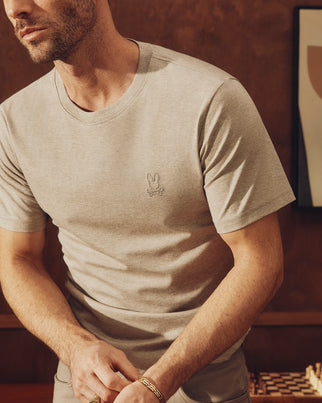 A man wearing a light gray Psycho Bunny Mens James Premium Crew Neck Tee (B6U952D200), made from Pima cotton and featuring a small embroidered logo on the chest, leans over a chessboard against a brown wall with framed artwork in the background.