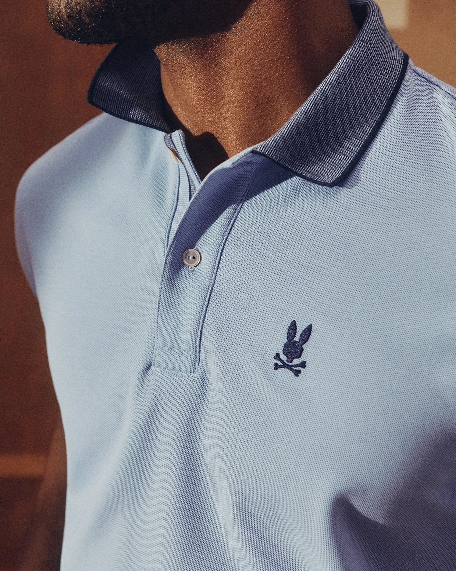 A person wears the Psycho Bunny MENS JONAS MICRO STRIPE COLLAR PIQUE POLO in light blue with a dark blue collar, showcasing an embroidered rabbit and bones logo on the left chest. Soft shadows play on the micro-striped fabric under gentle lighting.