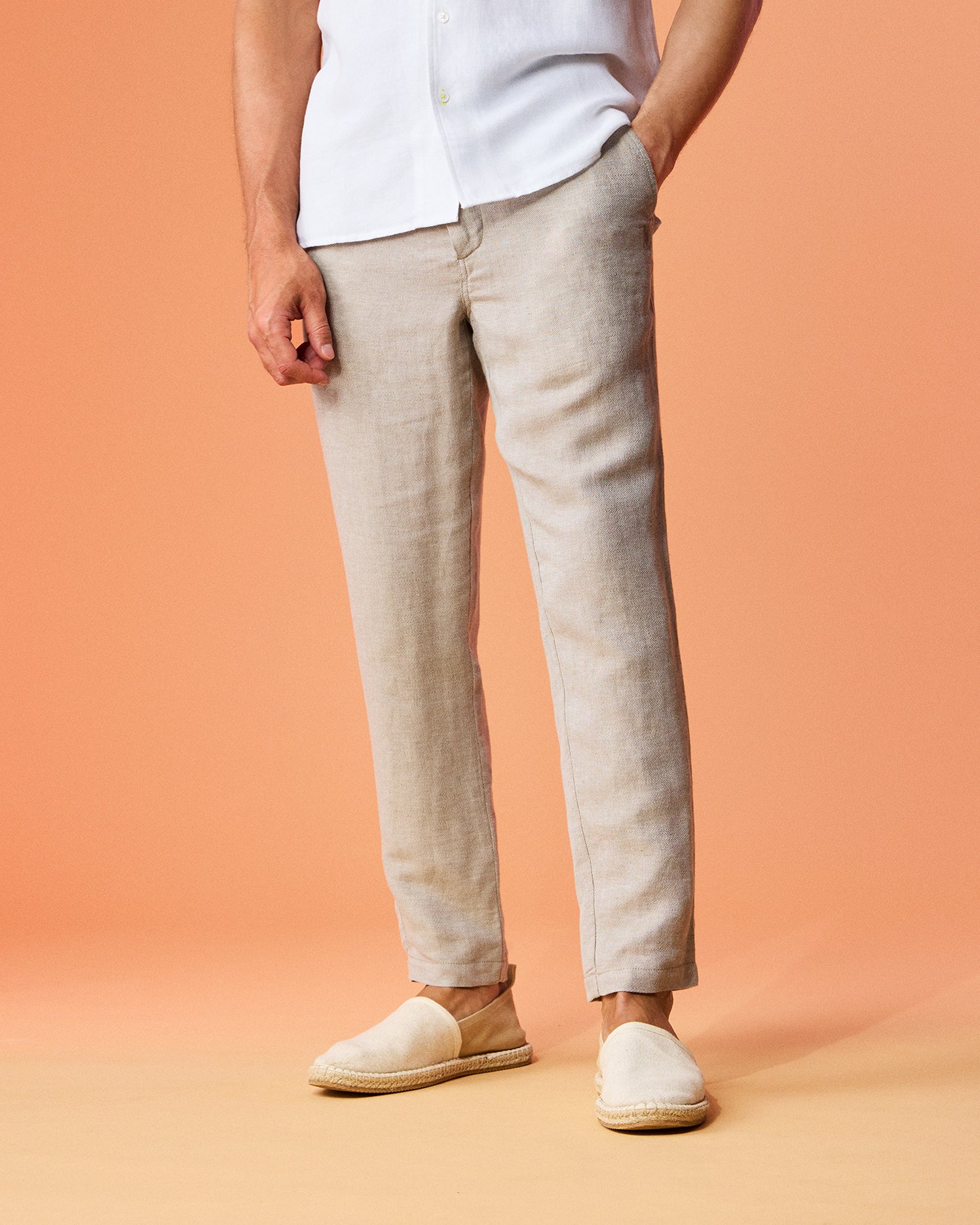 A person wearing the Psycho Bunny MENS FATE LINEN DRAWSTRING PANT - B6P471C200 and white casual shoes stands against a peach background, with a hand casually placed in the pocket. Only the lower torso and legs are visible.