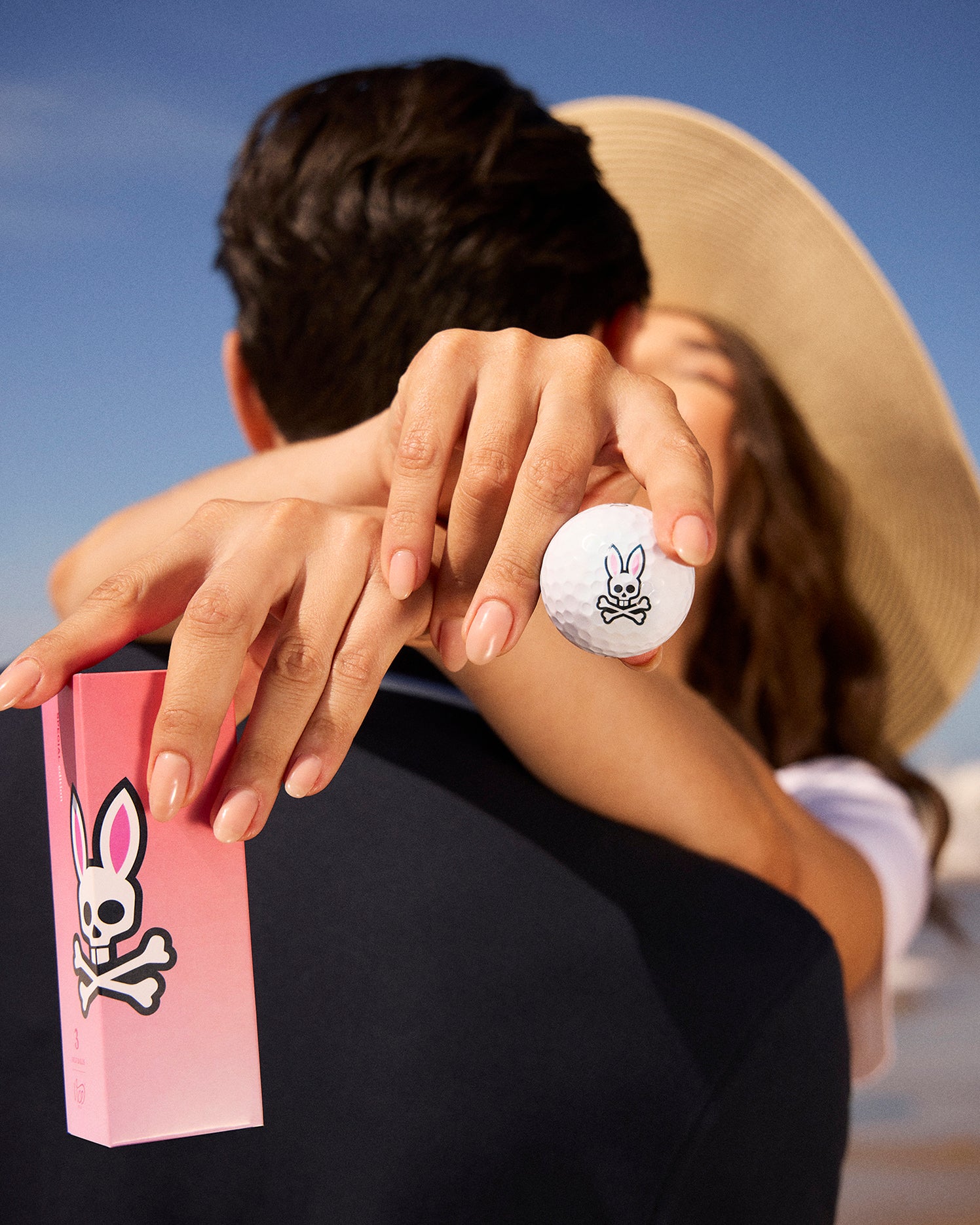 A woman with a wide-brim hat embraces a man, holding a pink box and a sleeve of 3 PACK GOLF BALLS - B6A565C200 featuring the Psycho Bunny logo. The background boasts a clear blue sky while the man's back is towards the camera, subtly showcasing gear designed to minimize driver spin.
