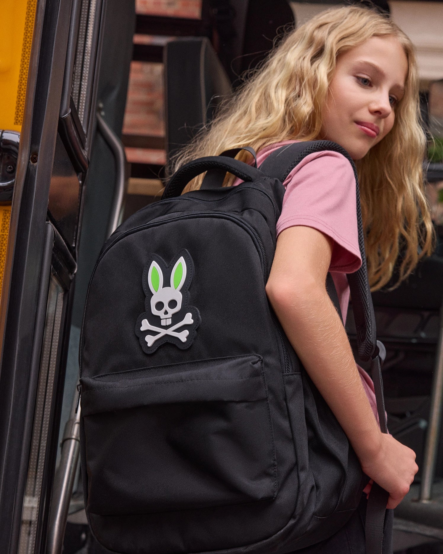 A young blonde girl wearing a pink t-shirt is stepping onto a school bus. She carries the Psycho Bunny KIDS BACKPACK - B0A175D200, featuring adjustable shoulder straps and a distinctive skull-and-crossbones design with bunny ears in green and white. The bus doors and interior are visible in the background.