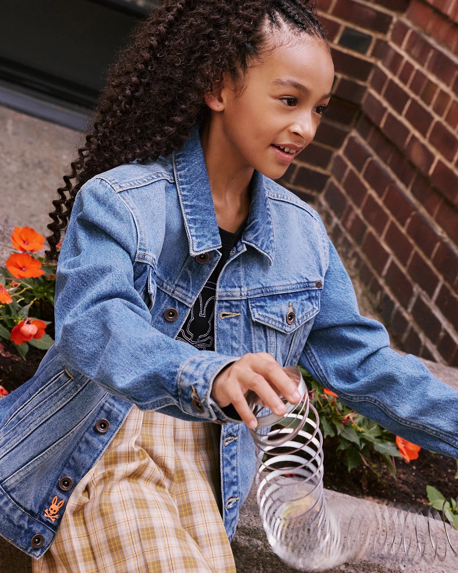 A young girl with curly hair wearing a KIDS PATHE DENIM JACKET - B0J741X1CO by Psycho Bunny and checkered pants plays with a silver slinky. She is outdoors, sitting near a brick wall decorated with vibrant orange flowers. The denim jacket, made from upcycled cotton, adds to her joyful and engaged demeanor.