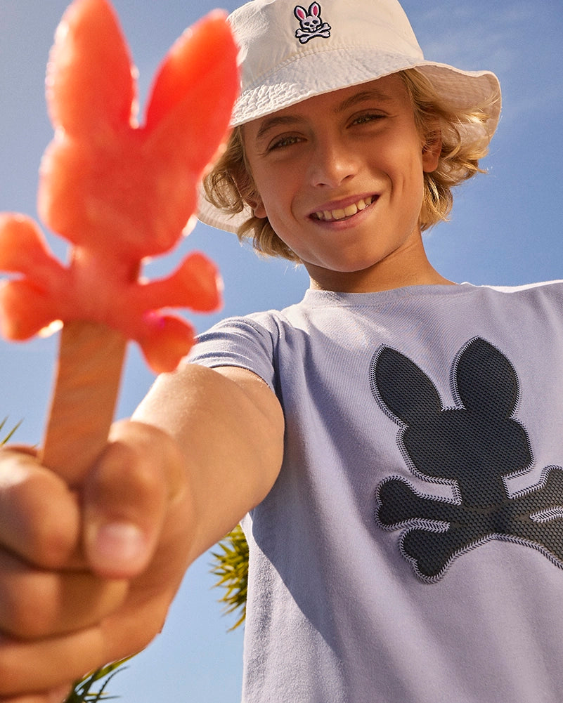 A smiling young person holds a pink bunny-shaped candy in the foreground. They are wearing a KIDS RIVIERA GRAPHIC TEE - B0U639C200 by Psycho Bunny with a black bunny and crossbones design and a white bucket hat featuring the same bunny logo. The background shows a clear blue sky and some greenery.