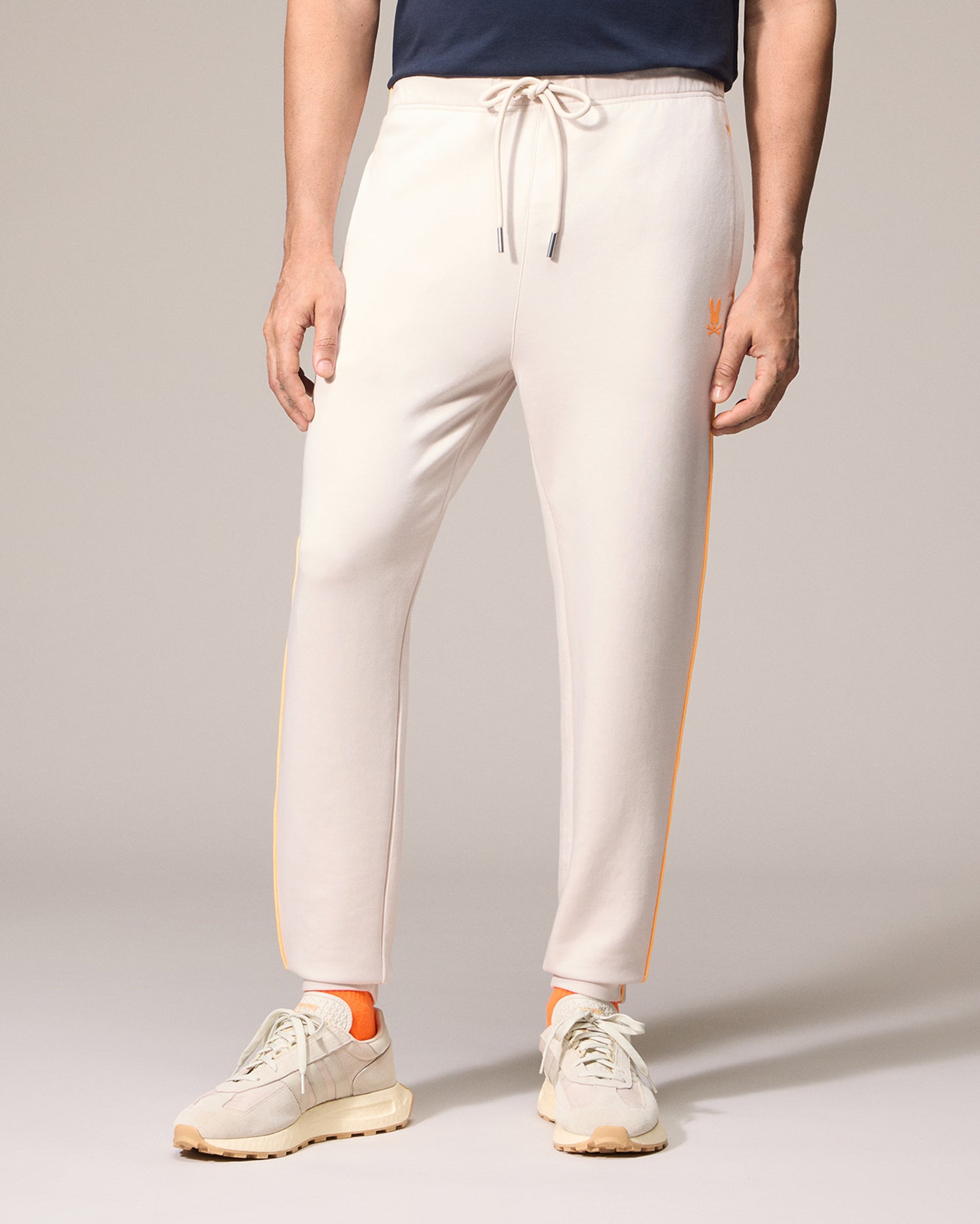 A man wearing cream-colored men's Psycho Bunny Leon sweatpants with an orange stripe down the side, paired with cream and orange sneakers, standing against a neutral background.