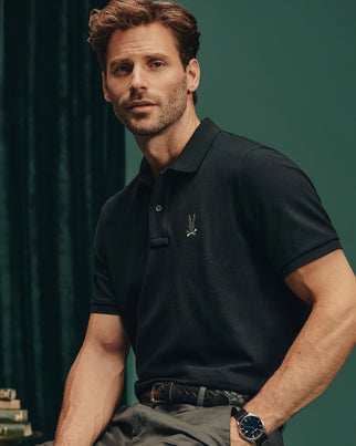 A man with short hair and a beard stands against a dark green background, wearing gray pants and accessorized with a watch. He sports the Psycho Bunny Men's Alexander Pique Polo - B6K101E200 in black, which features a small embroidered design on the left side, offering durable breathability.