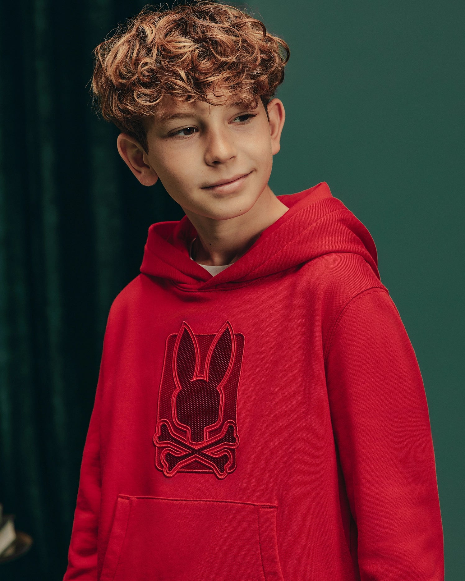 A young boy with curly hair wears the Psycho Bunny KIDS PIERCE ESSENTIAL FRENCH TERRY HOODIE - B0H135E200, showcasing a rabbit skull design, against a dark green background. He glances to the side with a slight smile.