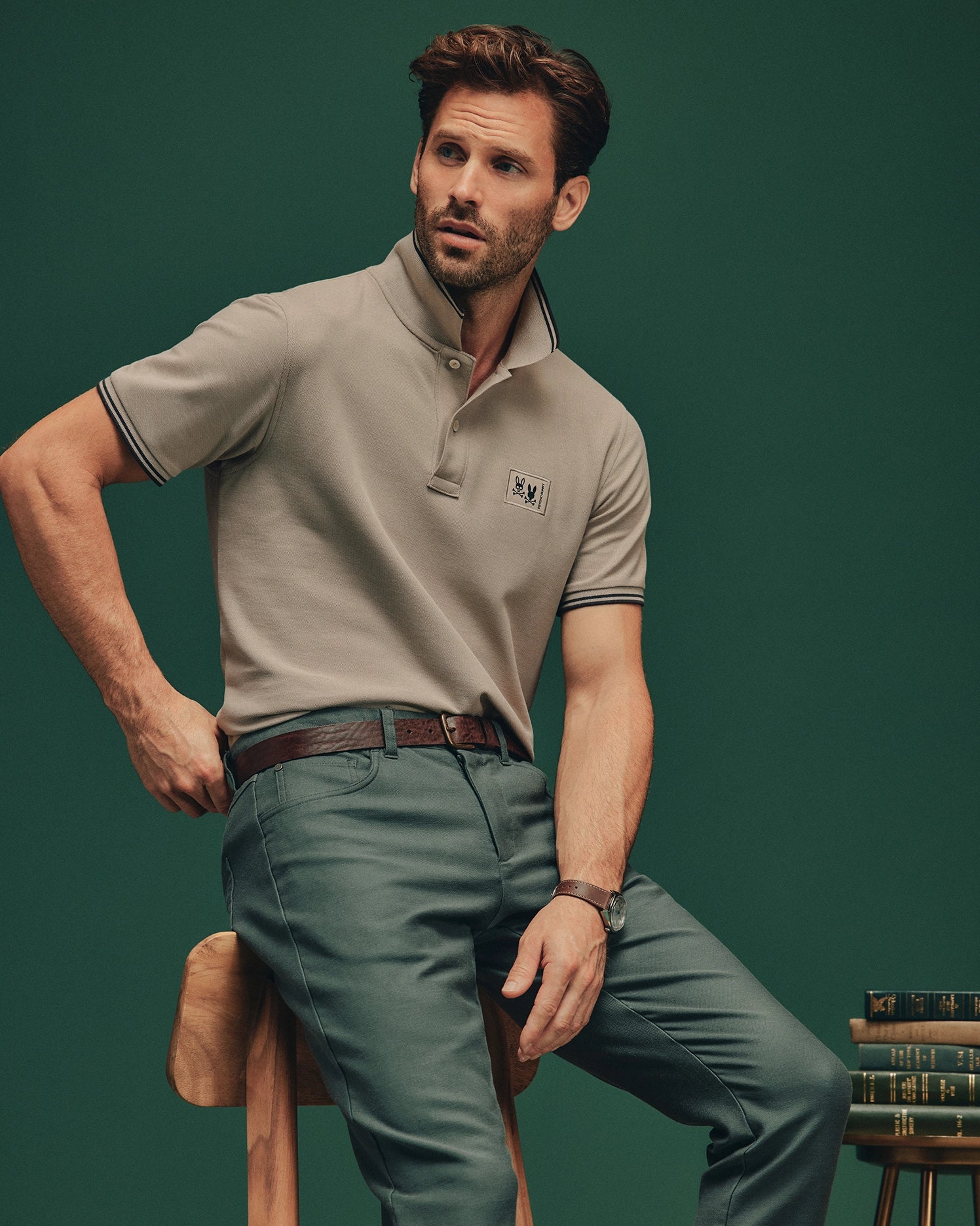 A bearded man is seated on a wooden stool in front of a dark green backdrop. He is dressed in a light gray Psycho Bunny Men's Monroe Pique Polo (B6K964D200), cut in a regular fit, complemented by green pants and accessorized with a brown belt and wristwatch. Nearby, there is a small stack of books.