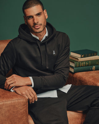 A man wearing a Psycho Bunny Men's Levy Essential Fleece Hoodie (Style B6H859D200), made from 100% cotton French terry, relaxes comfortably on a leather sofa. He's sporting a casual layered look with a slight smile, while stacks of books with green and gold spines add depth to the dark green backdrop behind him.