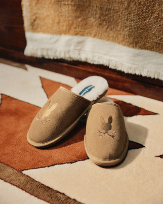 Resting on a geometric patterned rug is a pair of Psycho Bunny's MENS SUEDE SLIPPERS, crafted from cow suede leather with genuine shearling lining. Each slipper features the distinctive embroidered rabbit over crossbones design, with a fringed fabric in the background.