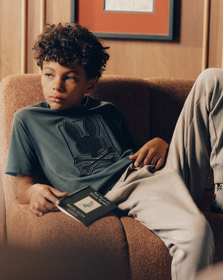 A boy with curly hair sits on a brown couch, wearing a dark shirt from the Psycho Bunny brand featuring an embroidered bunny logo, paired with light pants made from Kids' Pima Cotton. He holds a book in his right hand and gazes thoughtfully into the distance. A framed picture hangs on the wooden wall behind him.