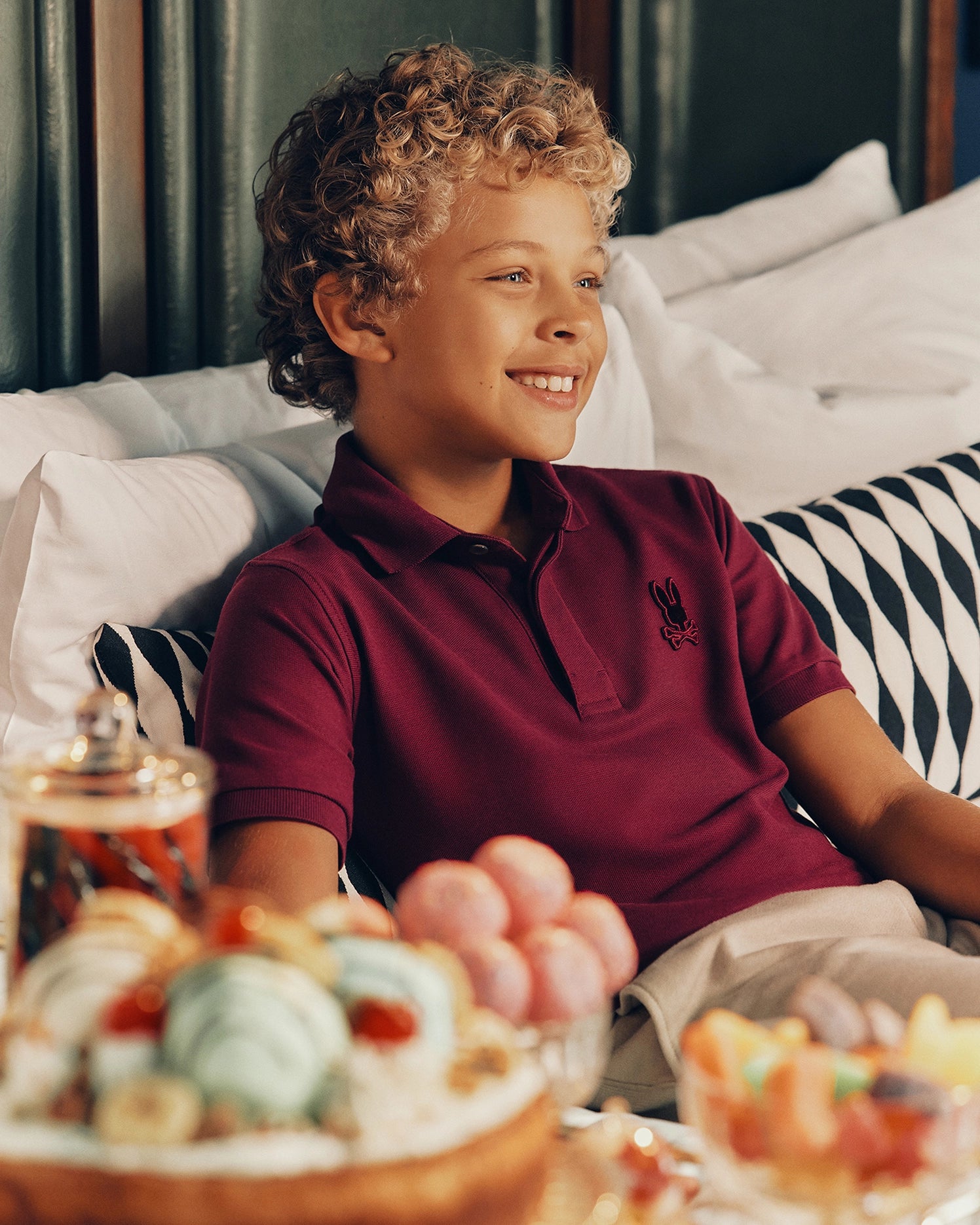 A cheerful child with curly hair, dressed in a stylish KIDS BRANDSON PIQUE POLO by Psycho Bunny, sits on a bed adorned with black and white striped pillows. In front, there are plates filled with vibrant macarons and pastries, creating a cozy and inviting atmosphere.