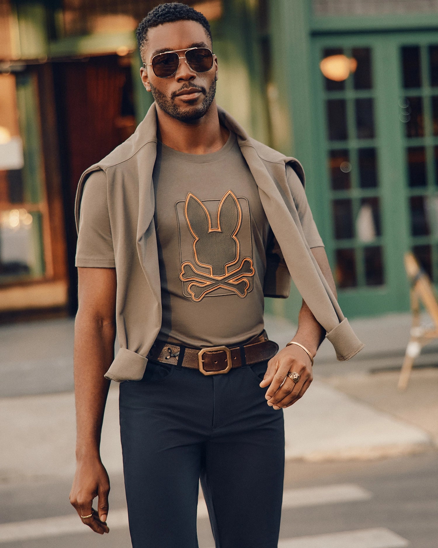 A stylish individual wearing sunglasses and the Psycho Bunny MENS PIERCE EMBROIDERED GRAPHIC TEE - B6U115E200, featuring bunny embroidery and a crossbones design, confidently walks down the street with a lightweight jacket draped over their shoulders. In the background stands a building with large windows and a green door.