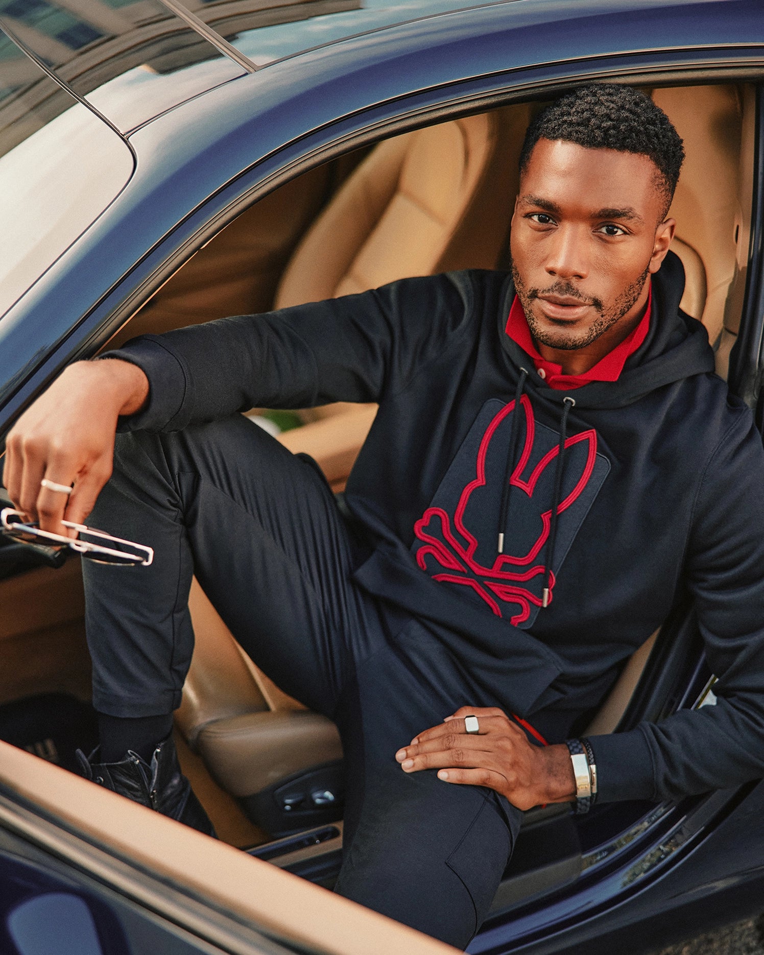 A man sits confidently in a car with the door open, wearing a stylish Psycho Bunny MENS PIERCE ESSENTIAL FRENCH TERRY HOODIE - B6H135E200, black pants, and holding sunglasses. A watch graces his wrist and rings adorn his fingers as he gazes at the camera.