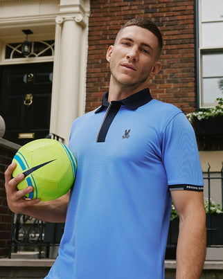 A young man stands outside, holding a yellow and blue soccer ball in his right hand. He is wearing a Psycho Bunny Mens Edmonds Sport Polo Shirt - B6K906D200, made from tech pique fabric, featuring a small logo on the chest and sporting a short haircut. The background features a brick building with a black door and the number 2 above it.