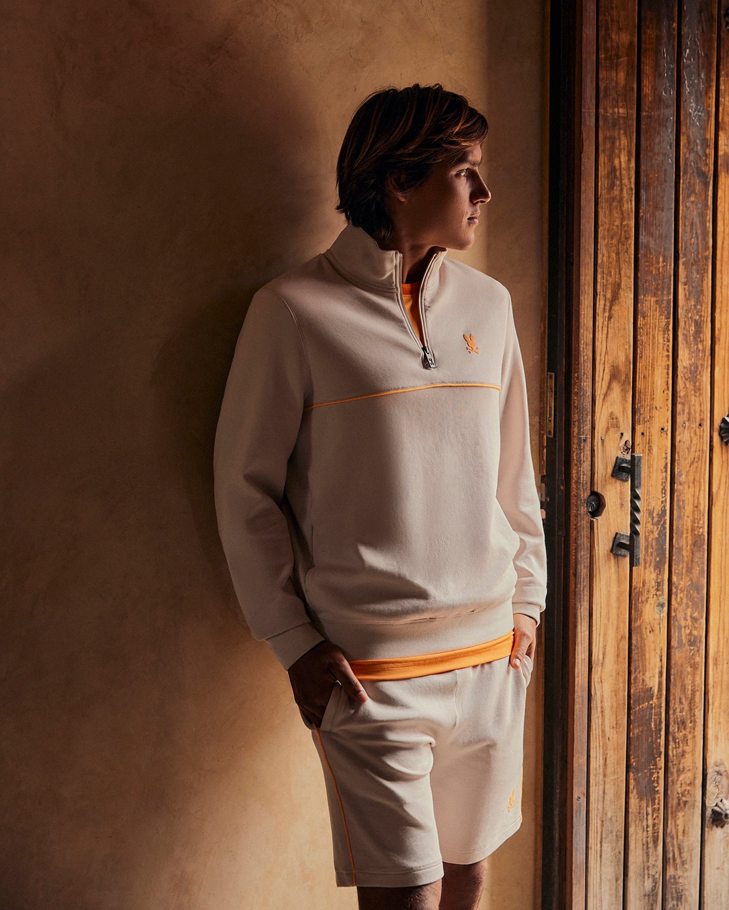 A person with short hair stands indoors, leaning against a beige wall near a wooden door. They are wearing a Psycho Bunny MENS LEON HALF ZIP SWEATSHIRT - B6S204B200 with orange accents and matching beige shorts with orange trim, hands in pockets, looking to the side.