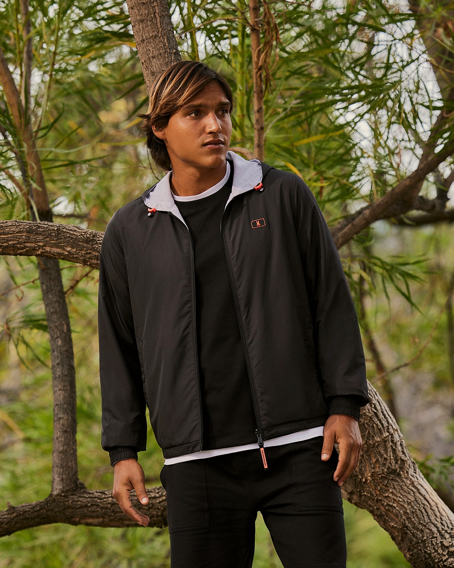 A person with long hair stands outdoors near tree branches, wearing a black water-repellent Psycho Bunny MENS HALFMOON REVERSIBLE JACKET - B6H169B200 over a white and black shirt. The background shows green foliage, highlighting a natural setting. The person is posing with one arm resting on a tree branch.