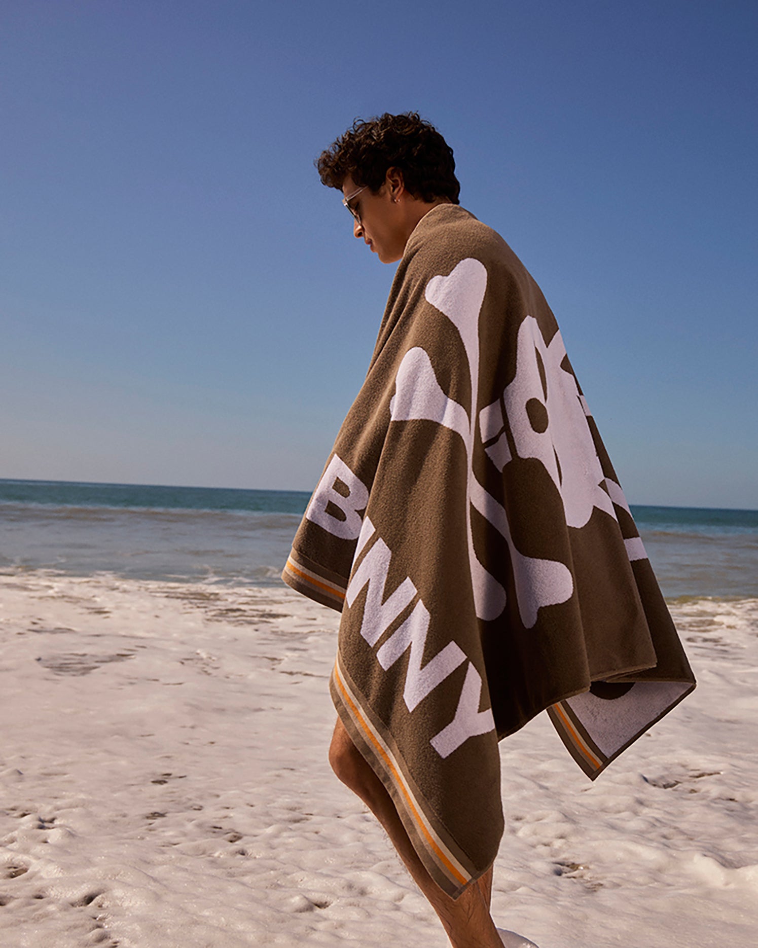 A person with short dark hair stands on a sunny beach, wrapped in the Psycho Bunny BEACH TOWEL - B6A393B200 featuring a large white skull and crossbones design. The ocean and blue sky are visible in the background. The jacquard-knit Bunny logo and text adorn the striped border at the bottom of the towel.