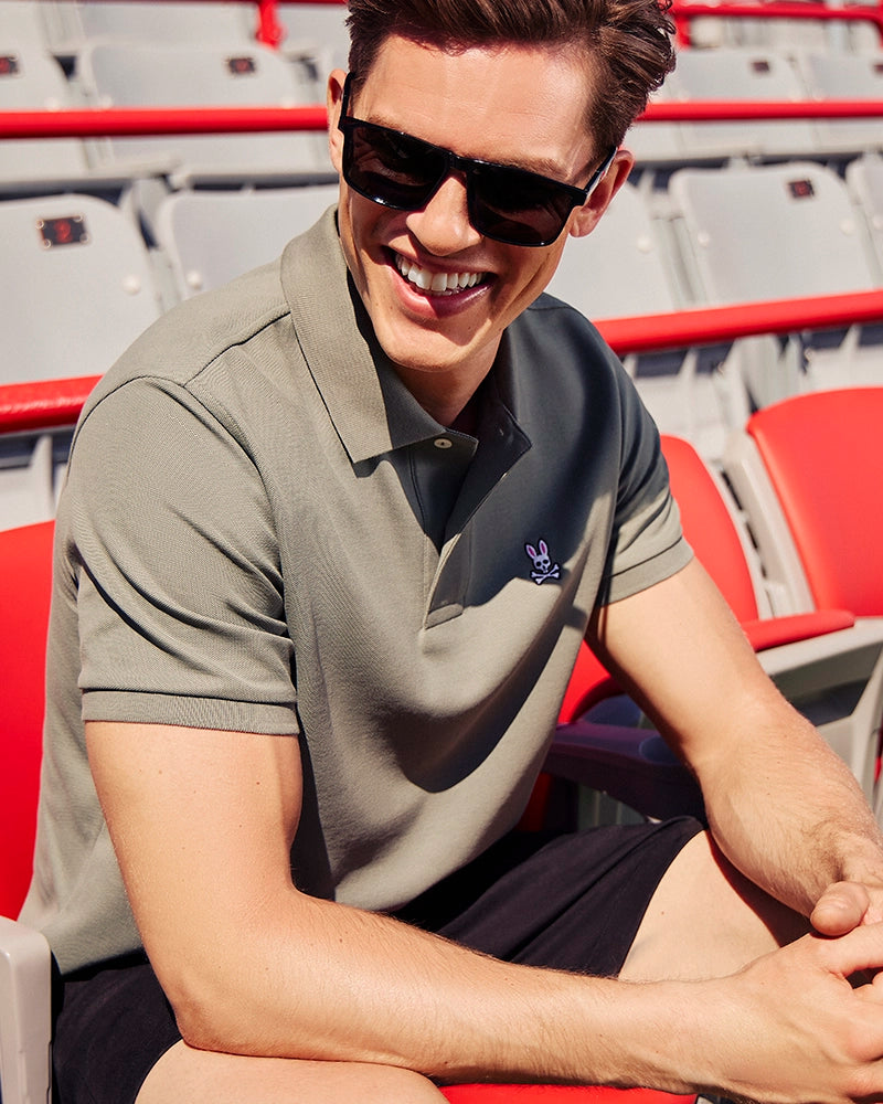 A man with short brown hair and sunglasses sits smiling in a stadium seat. He is wearing a green Psycho Bunny MENS CLASSIC PIQUE POLO SHIRT - B6K001B200 made of Pima cotton, featuring a small embroidered Bunny logo, and black shorts. The background shows rows of red and grey seats.
