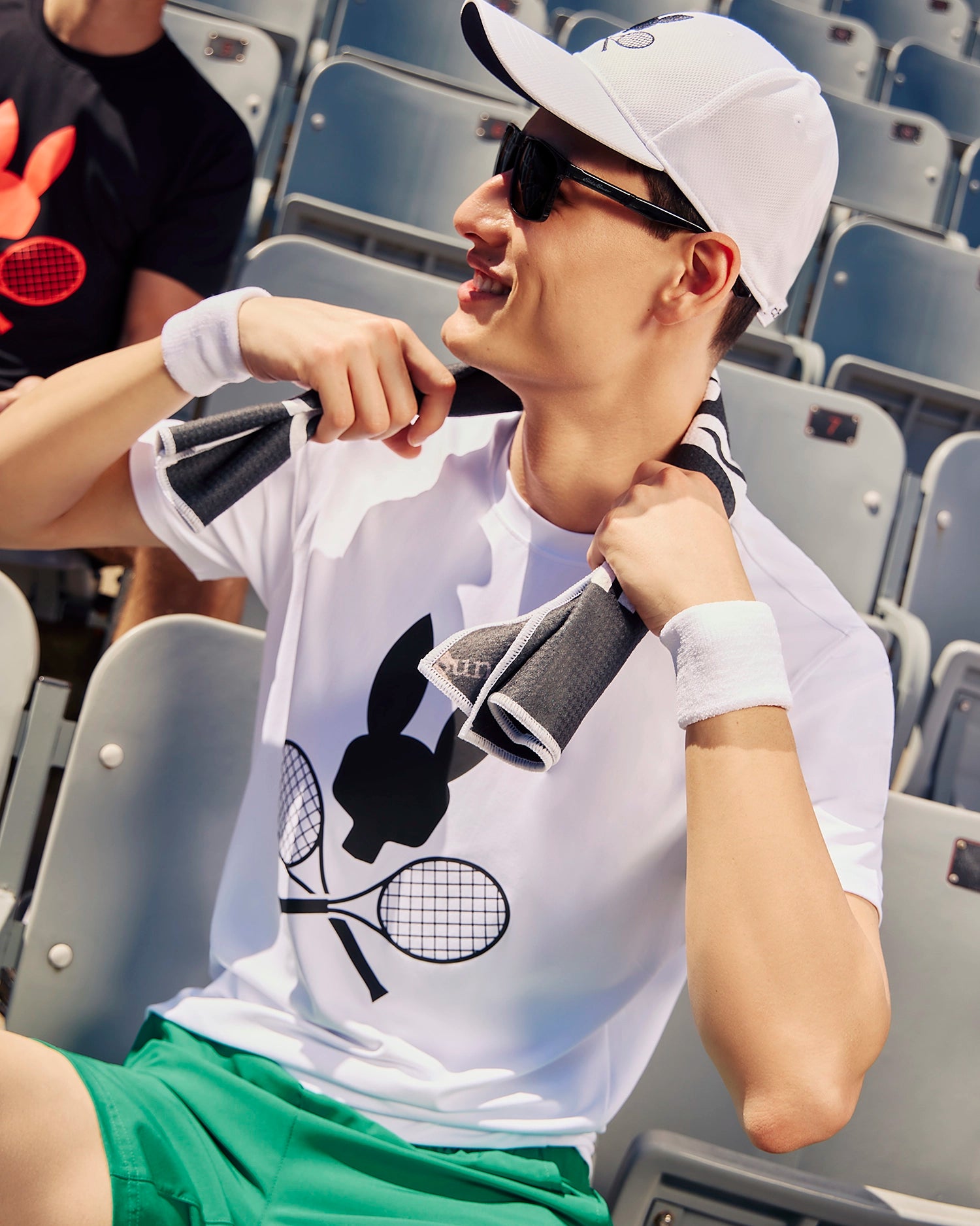 A person wearing a white shirt with a tennis-inspired Psycho Bunny logo, white cap, sunglasses, and green shorts sits in stadium bleachers. The individual is holding a towel around their neck and wearing wristbands, appearing relaxed and happy. Another person is seated nearby in the background.