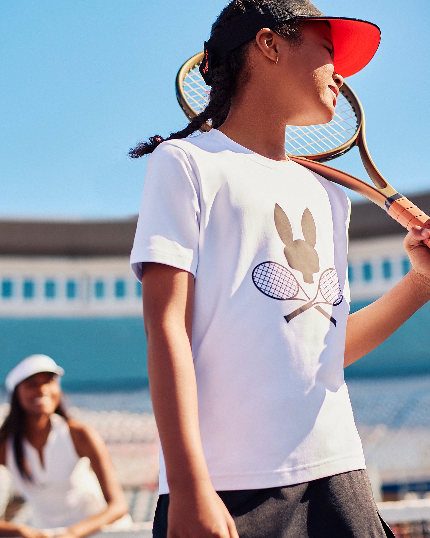 A young girl wearing a Psycho Bunny KIDS COURTSIDE GRAPHIC TEE - B0U681C200 featuring a rabbit and crossed tennis rackets, paired with a red visor, holds a racket over her shoulder. Another person in a white cap and outfit stands in the blurred background of the sunny tennis court.